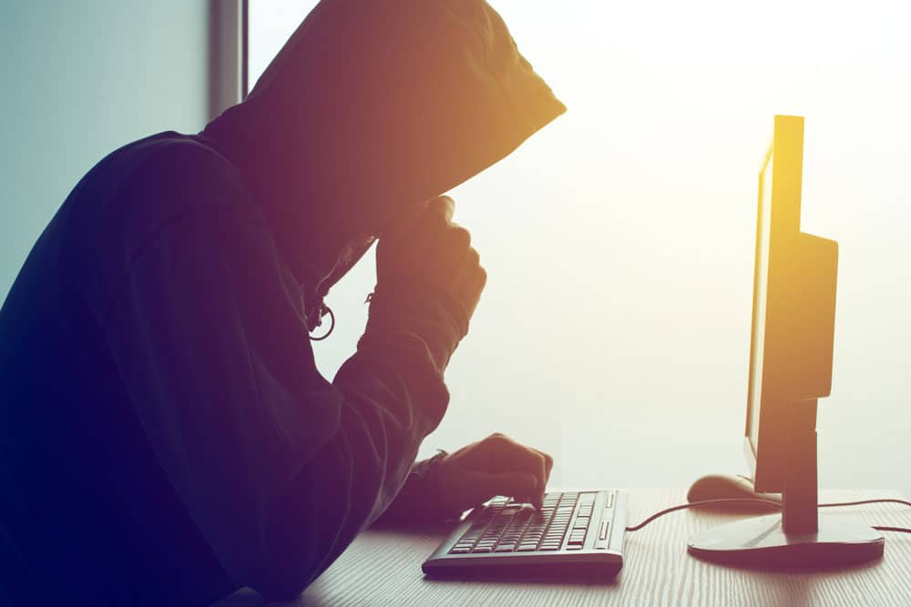 hacker wearing hoodie sitting at desk with computer