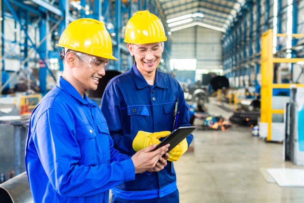 Two male warehouse workers in blue utility suits and yellow hard hats holding a tablet and smiling