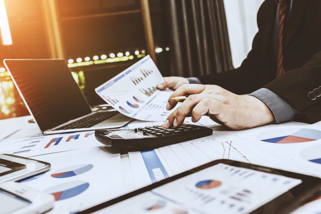 businessman sitting at desk with laptop and calculator with paperwork and graphs spread out on desk